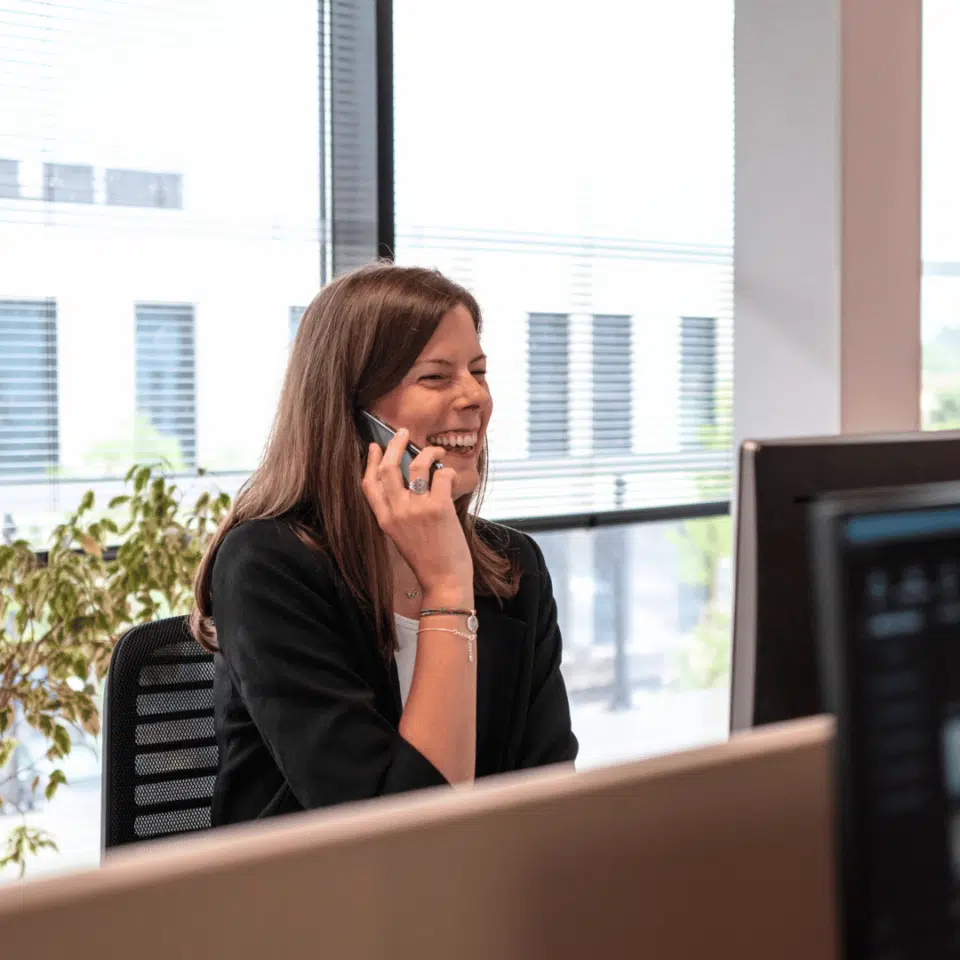 Femme souriante assise à un bureau dans un bureau moderne et lumineux, parlant au téléphone.